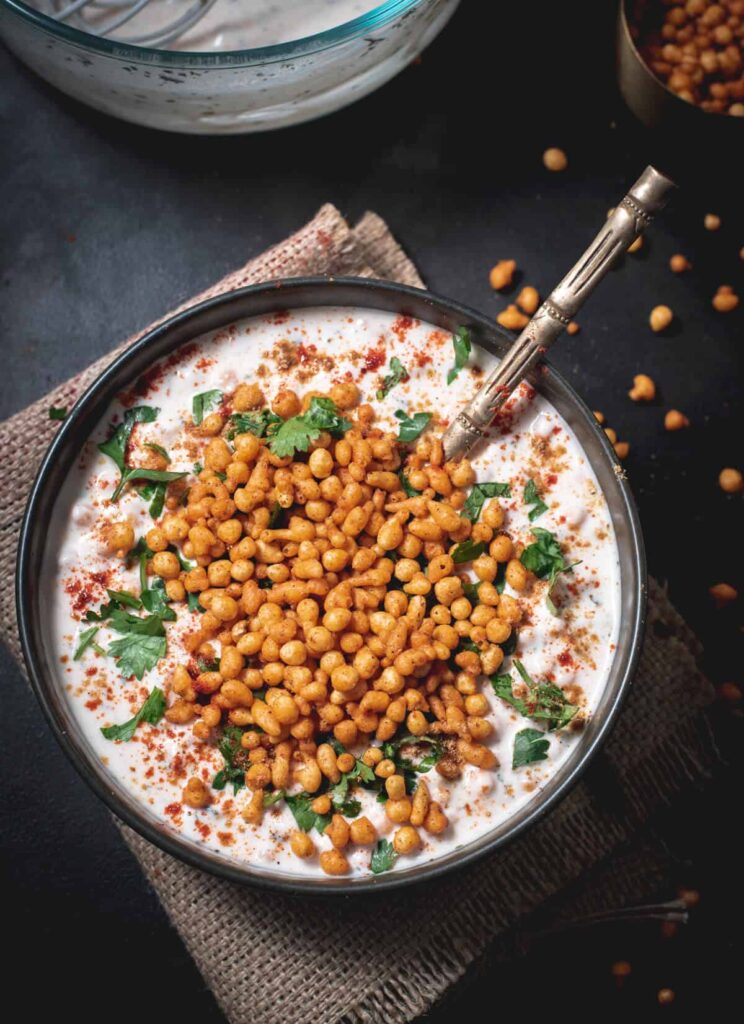 An overshot of boondi raita garnished with cilantro and served in a black bowl with a golden spoon.