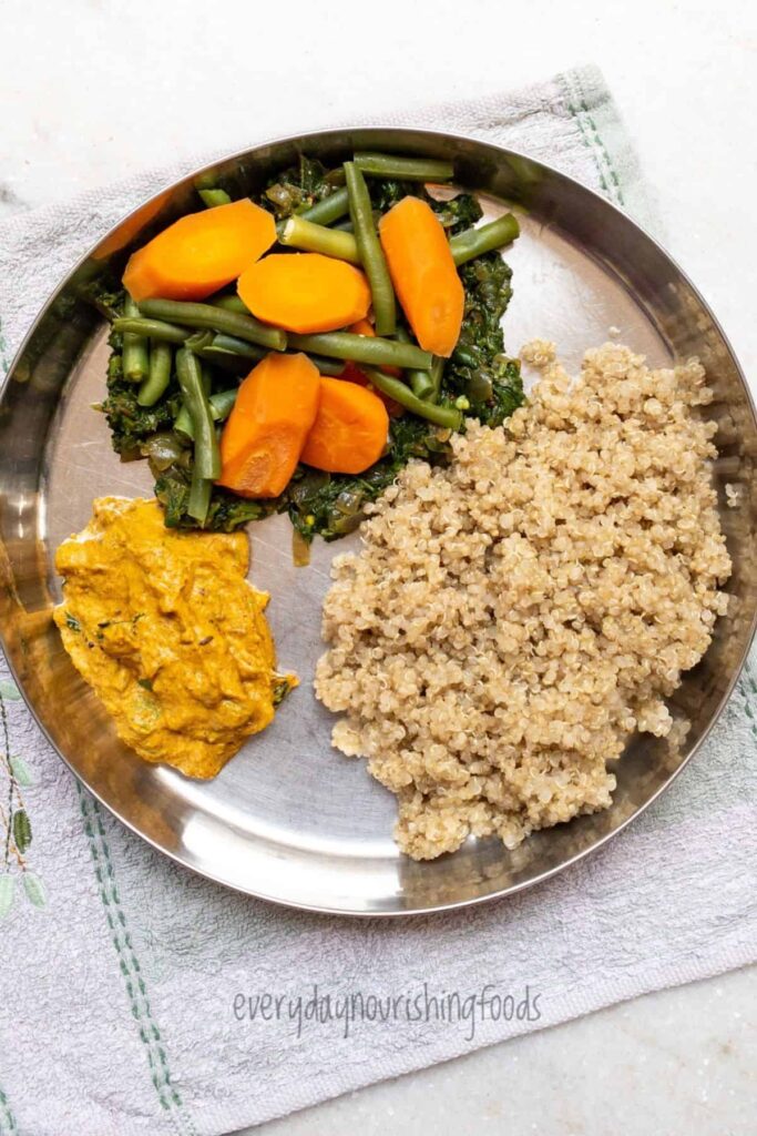 A balanced meal served on a stainless steel plate featuring steamed carrots and green beans, saag, tomato chutney, and a side of quinoa, with