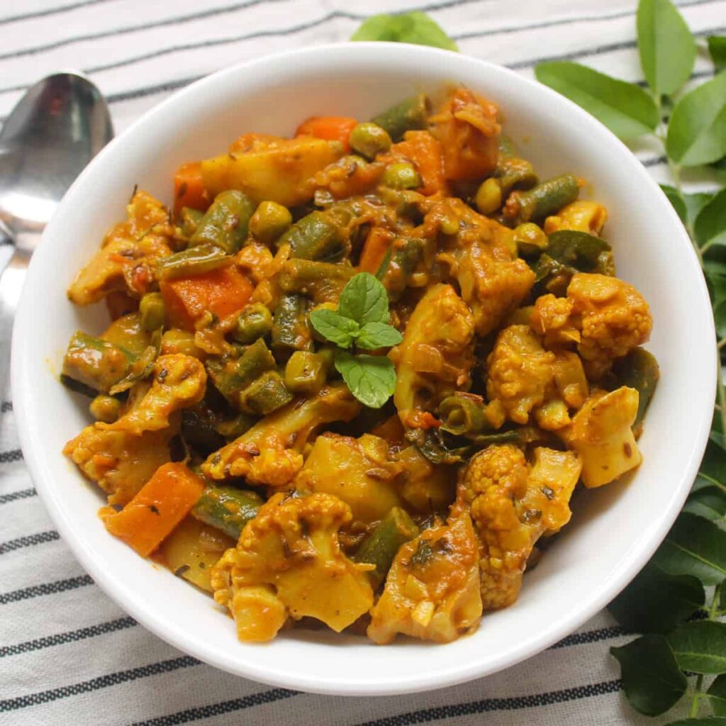 A bowl of Indian vegetable curry garnished with fresh mint, served with a side of curry leaves on a striped napkin.