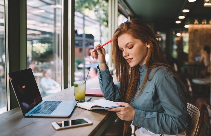 Side view of young female freelancer with pen touching hair and looking down at notebook while sitting at desk with laptop smartphone lemonade glass and worried about work progress in blurred cafe