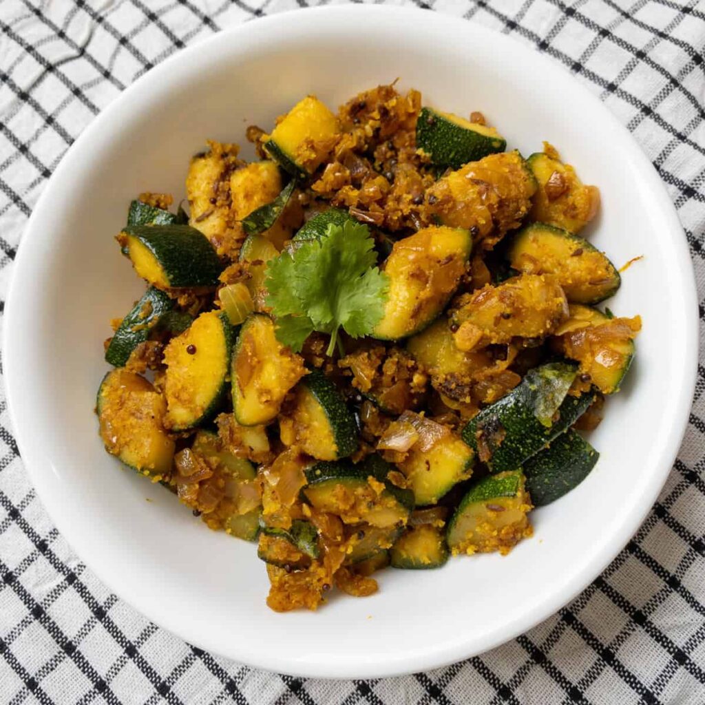 Bowl of dry-style Indian zucchini curry garnished with cilantro, served on a black and white checkered cloth.