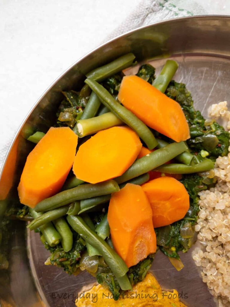 Close-up of steamed green beans and sliced carrots atop saag on a stainless steel plate, next to quinoa, with 'Everyday Nourishing Foods' watermark.